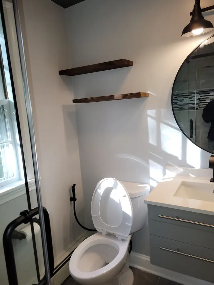 Bathroom corner with toilet, walnut floating shelves, round black mirror and glass shower door
