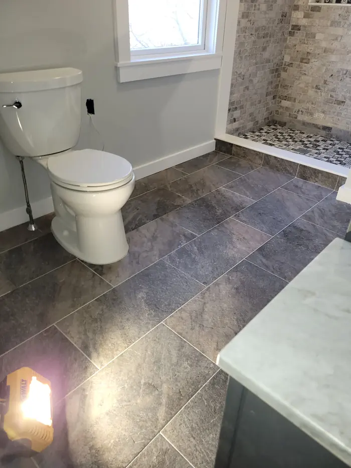 Dark grey plank-style floor tile in a remodeled bathroom with white toilet and stone shower beyond