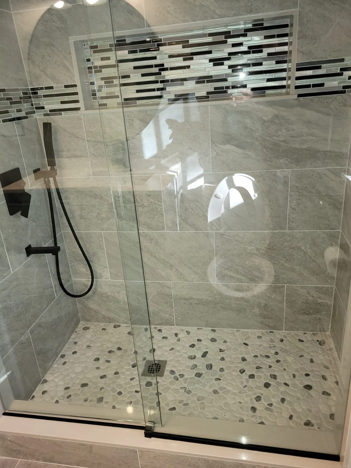 Frameless glass shower door over a pebble tile shower floor with matte black handheld shower and grey porcelain walls