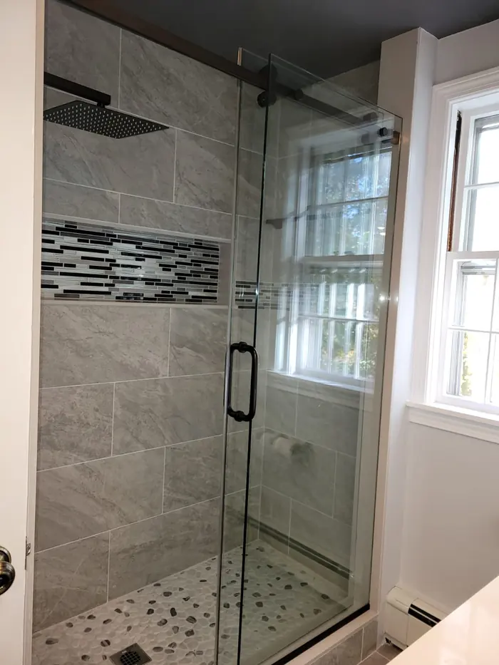 Glass shower enclosure with grey porcelain tile and mosaic band next to a window in a remodeled bathroom