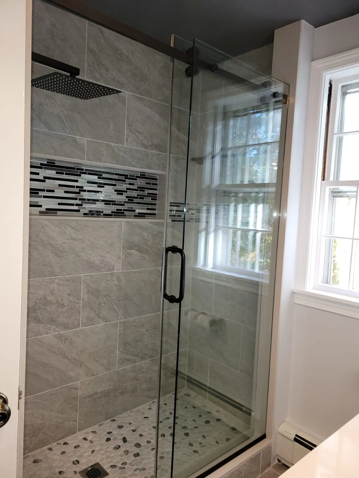 Glass shower enclosure with grey porcelain tile and mosaic band next to a window in a remodeled bathroom