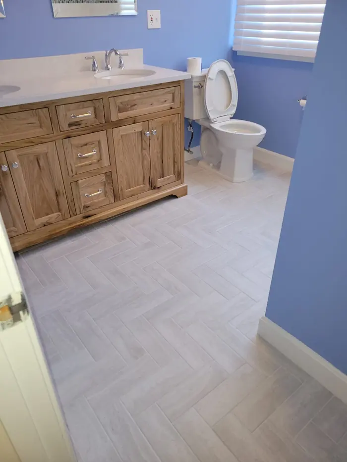 Light grey herringbone-pattern tile floor running past a rustic wood vanity in a blue bathroom